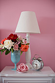 Vase of roses, table lamp and old-fashioned alarm clock on bedside cabinet against pink wall