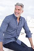 A grey-haired man on a terrace wearing a blue shirt and jeans