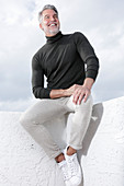 A grey-haired man on a terrace wearing a grey turtleneck jumper and light trousers