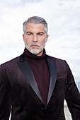 A grey-haired man on a terrace wearing a dark-red turtleneck jumper and a jacket