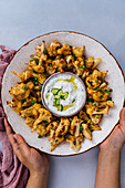 Hands holding roasted spicy cauliflower wings in a ceramic bowl served with a small bowl of yogurt cucumber dip