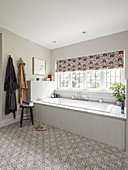 Bathtub with white-tiled surround below window with patterned Roman blind