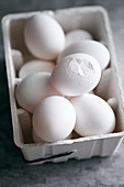 White chicken eggs in egg carton, one with a slightly broken shell