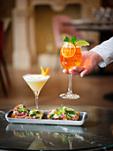 Hands holding yellow and orange cocktail decorated with mint and lime and a dish of roast beef