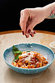 Hand sprinkling with grated cheese on noodles with tomato sauce and basil leaves in blue bowl