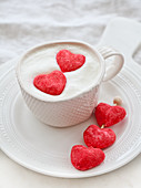 A cup of white hot chocolate with whipped cream and two pink heart marshmallows