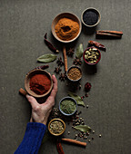 hand holding pot with red paprika powder over gray table with set of assorted aromatic spices