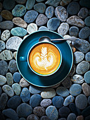A fresh coffee in a blue cup and saucer with fern pattern in the foam crema froth, against a stone background