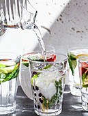 Various glasses of water filled with fruit, mint, and ginger with water being poured from a pitcher into one glass