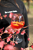 A hand holding a glass of Aperol Spritz with a sprig of blossom in the foreground