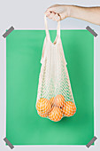 Hand carrying net bag with ripe oranges against green rectangle during zero waste shopping