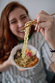 Cheerful redhead female laughing and picking noodles from bowl of tasty ramen while sitting on couch at home
