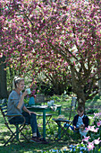 Sitzgruppe unter Zierapfelbaum 'Paul Hauber', Frau mit Kaffeetasse, Junge sitzt und liest, Hund Zula