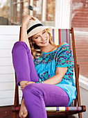 Young blonde woman in hat, blue tunic blouse and purple trousers in deck chair