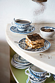 Tiramisu slice on a blue plate on marble with coffee and chocolate powder falling