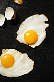Two fried quail eggs on black cooktop surface with cracked egg shells to the side.