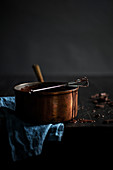 Melted dark chocolate in copper pot with drops going down side. Small metal whisk with chocolate, blue linen on a black wood surface. Dark, moody setting.
