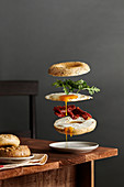 A bagel breakfast sandwich with ingredients floating. Rustic wood table setting against a gray wall.