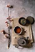 Tea drinking wabi sabi japanese style dark clay cups and teapot on cloth napkin with blooming cherry branches. Grey texture concrete background. Flat lay, space
