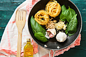 Bottle of olive oil, frying pan with raw tagliatelle rolls, garlic with green basil leaves and crispy pine nuts