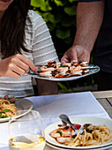 Platter of shrimp skewers being passed an an outdoor table with pasta and white wine