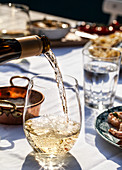 White wine being poured into a glass, on an outdoor table with pasta cacio e pepe (cheese and pepper), basil and shrimp skewers