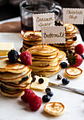 Stacks of mini pancakes with signs, with fruit and chocolate chips, syrup and butter, on a marble board