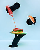 Hand in black gloves holding plate with burger and spilling ketchup on patty against blue background