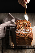 Pouring white sugar icing over homemade banana bread with nuts placed on metal grid on wooden table