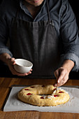 Crop chef in black apron greasing unbaked round bread with egg yolk topped with cherry while standing at wooden table