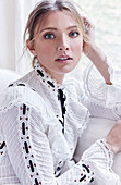 A blonde woman wearing a fancy white blouse decorated with black ribbon sitting in front of a window