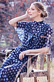 A blonde woman wearing a blue, polka dot dress sitting in a garden