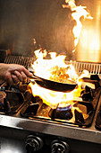 Chef tossing frying shrimps in flambe above gas stove with burning flame while cooking in restaurant