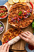 Hands rolling up a lahmacun on a wooden board with some more lahmacun portions and condiments