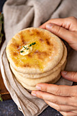 Hands holding a stack of Turkish flatbread called bazlama topped with butter and thyme on a wooden board.