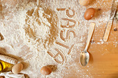 Top view of inscription Pasta written with finger on wooden kitchen table covered by flour with olive oil eggs and kitchen utensil locating on table