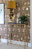 A lamp on a filigree console table against a wall with floral wallpaper