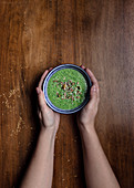 Hände halten grüne Smoothie Bowl mit Kürbiskernen und Sesam auf Holztisch