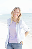 A blonde woman by the sea wearing a purple t-shirt and a blue-and-white striped shirt