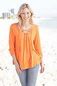 A blonde woman by the sea wearing an orange tunic and jeans