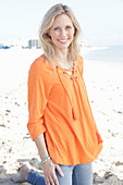 A blonde woman by the sea wearing an orange tunic and jeans