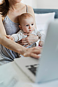 Mother holding baby daughter and working at laptop