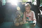 Girl with teddy bear using tablet in back seat of car