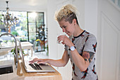 Woman drinking water and working at laptop in kitchen
