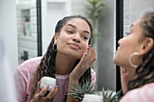 Beautiful woman applying moisturizer in bathroom mirror
