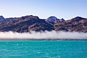 Fog below rugged mountains and turquoise ocean Greenland