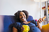 Laughing pregnant woman with tea and smart phone on sofa