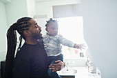 Father with long braids holding toddler son in kitchen