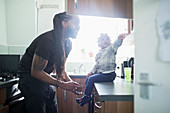 Happy father son putting shoes on toddler son in kitchen