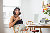 Portrait confident pregnant woman drinking tea at laptop